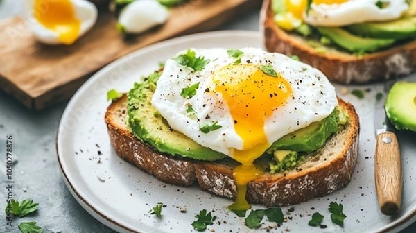 Fototapeta Avocado toast topped with a poached egg, with a wooden knife and a cutting board in the background.