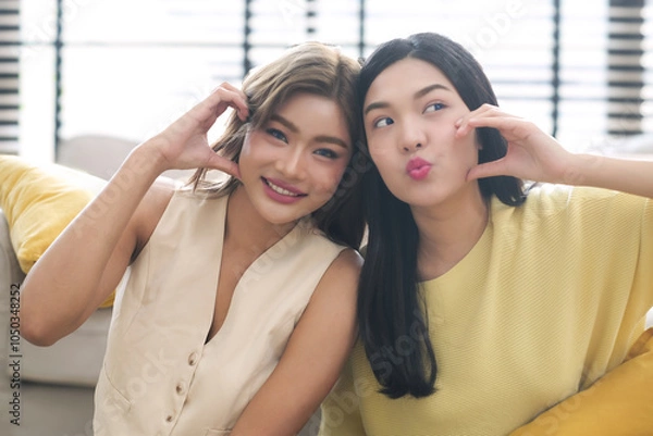 Obraz Portrait two young asian woman happy smile showing heart hands touch cheek gesture on sofa at home.