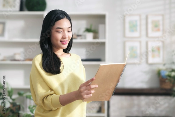 Obraz Smiling asian woman happily looks at a note book with names of customers ordering online in preparation for delivery.
