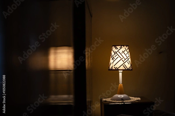 Fototapeta A close-up of a small table lamp on a nightstand in a dimly lit room. The lamp has a patterned fabric shade and casts a warm glow. The rest of the room is dark, with only the soft light from the lamp 