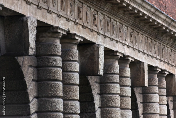 Fototapeta A grand colonnade with robust, textured stone pillars stands in quiet strength. The repetitive arches create a rhythmic architectural pattern against a high brick wall, surrounded by greenery.