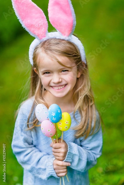 Obraz Beautiful smiling little girl with long blond hair wearing pink rabbit or bunny ears and blue knitted dress and holding bunch of painted colour eggs on sunny day in spring park. Easter celebrations