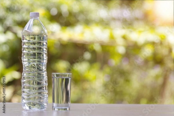Obraz A clear plastic water bottle and a glass of water are placed on a table against a blurred background of green leaves.