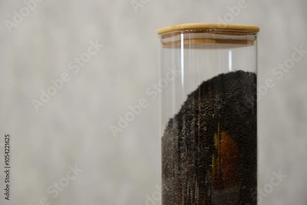 Fototapeta A clear glass container filled with black tea mixture sitting on a wooden table against a light textured background