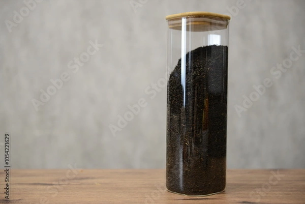 Fototapeta A clear glass container filled with black tea mixture sitting on a wooden table against a light textured background