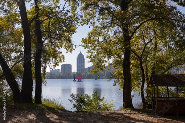 Obraz A serene view of a lake framed by trees, with red sailboats and a cityscape in the background under a clear sky