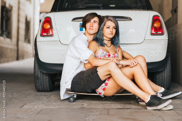 Fototapeta Couple hugging each other sitting on the skateboard desk in front of the car 