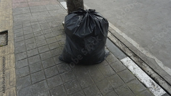 Fototapeta A black garbage bag that has been filled with garbage on the sidewalk during the day
