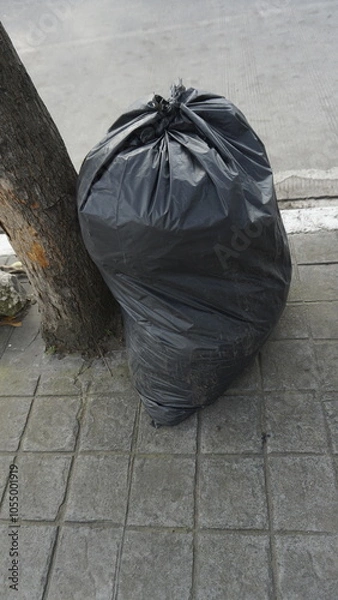 Fototapeta A black garbage bag that has been filled with garbage on the sidewalk during the day
