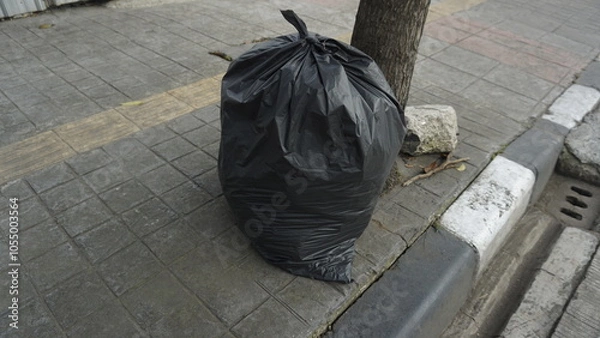 Fototapeta A black garbage bag that has been filled with garbage on the sidewalk during the day
