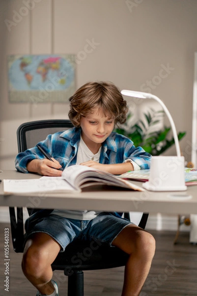 Fototapeta in the children's room a schoolboy in white sweater and a blue checkered shirt at a desk doing homework on his studies writing in a notebook
