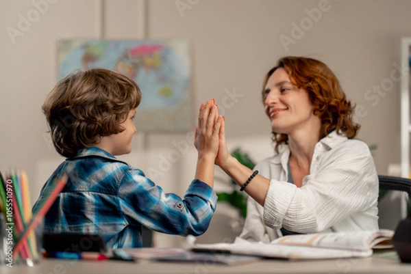 Fototapeta in the children's room schoolboy in a white sweater and a blue checkered shirt at a desk doing homework with his mother everything is done giving each other high fives