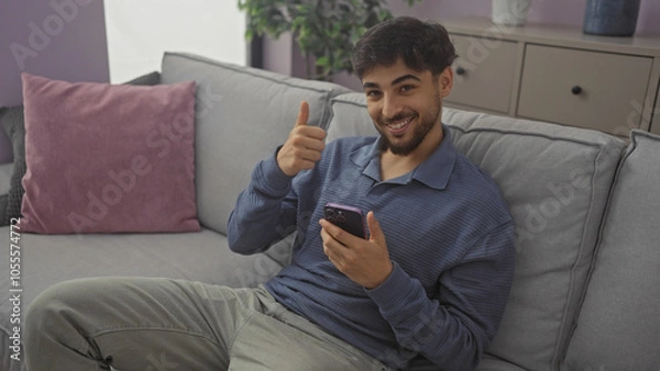 Fototapeta Handsome arab man with beard giving thumbs up while using smartphone on couch indoors.