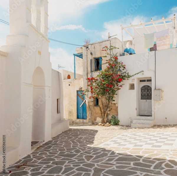 Fototapeta A beautiful corner of a traditional village in Paros, Greece, featuring whitewashed walls, a vibrant bougainvillea tree, and a characteristic blue door. 