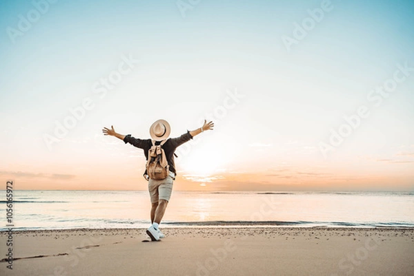 Fototapeta Happy man with backpack standing with arms up at the beach - Delightful tourist enjoying summer vacation by the seaside - Traveling life style and well being concept