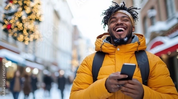 Fototapeta A joyful man in a yellow jacket holds a phone and laughs heartily while standing in a lively snow-covered street, against a backdrop of festive lights.
