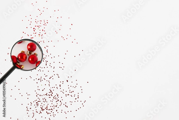 Obraz Looking through a magnifying glass on a red Christmas ball over a gliitter on a white background. Christmas time idea, holiday decoration concept. Copy space.