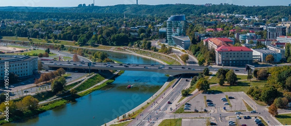 Fototapeta Aerial perspective of Vilnius, Lithuania, showing the Neris River, a bridge with vehicles, a boat, modern and historic buildings, and lush greenery.