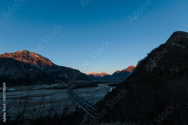 Fototapeta panoramica su un ambiente naturale nelle Alpi nel nord Italia, con un ampia valle attraversata dal fiume Tagliamento, all'ombra delle montagne, con cielo sereno, al tramonto