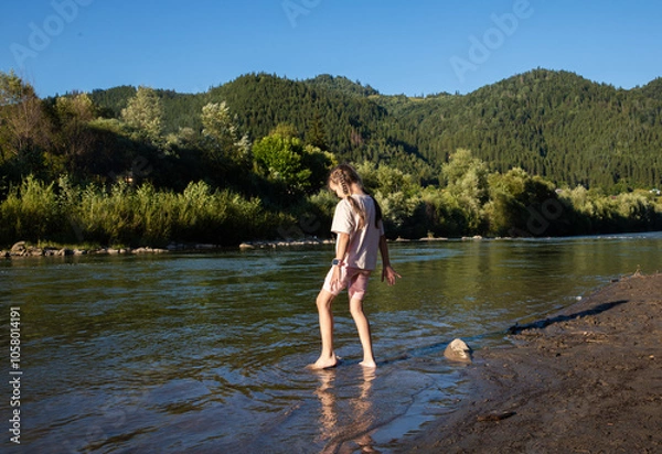 Obraz A girl stands in shallow water, feeling the cool river flow around her feet while sunlight filters through surrounding trees, creating a serene atmosphere filled with nature's beauty.