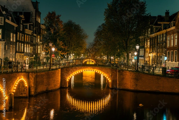 Obraz Amsterdam’s historic canal bridges illuminated at night. The Keizersgracht and Reguliersgracht intersection glows romantically, reflecting typical Dutch architecture and city lights in the water.