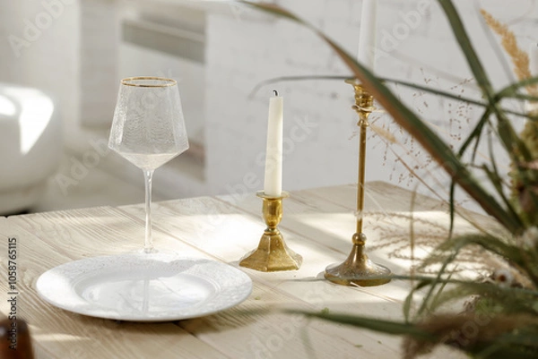 Fototapeta Elegant table setting with candle holders, white plate, and wine glass in natural sunlight