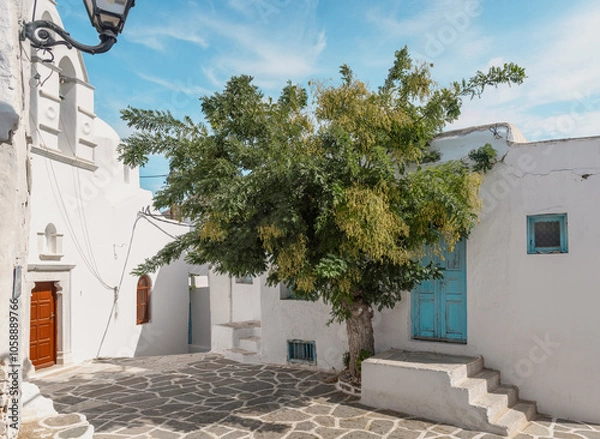 Obraz A serene, sunlit alley in Paros, Greece, featuring whitewashed walls, vibrant blue doors, and a lush tree. The Cycladic architecture and stone-paved path evoke the authentic ambiance of Greek life.