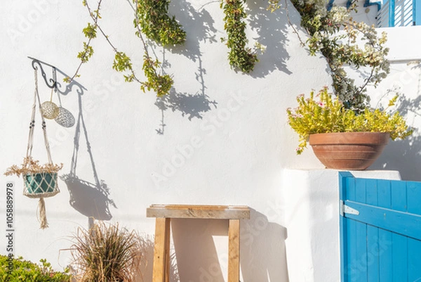 Obraz A serene outdoor space featuring a rustic wooden table, lush plants, and a striking blue door. The sun casts gentle shadows, enhancing the island's charm in Paros, Greece.