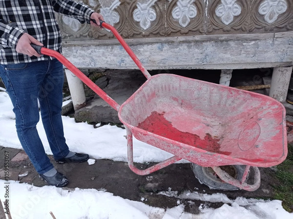 Fototapeta Man with snow cleaning tools - wheelbarrow and bucket, working utility, municipal community service. garden car in the country. the snow is melting and spring is coming. sunny spring day. close up.