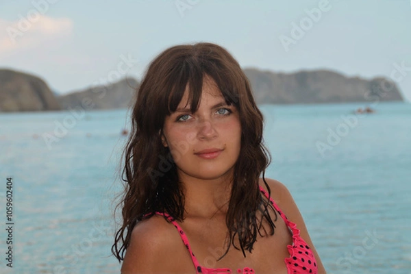 Fototapeta The face of a tanned girl with blue eyes. Girl against the backdrop of the sea coast. Beautiful girl with model appearance.