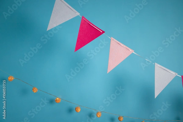 Fototapeta Close-up of festive triangle bunting in pink, red, and white colors alongside warm string lights against a bright blue background.