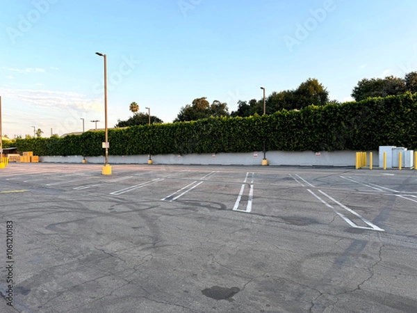 Obraz urban parking lot - city car park - empty parking spaces, wall with hedges, bollards, street lights, trees in the background