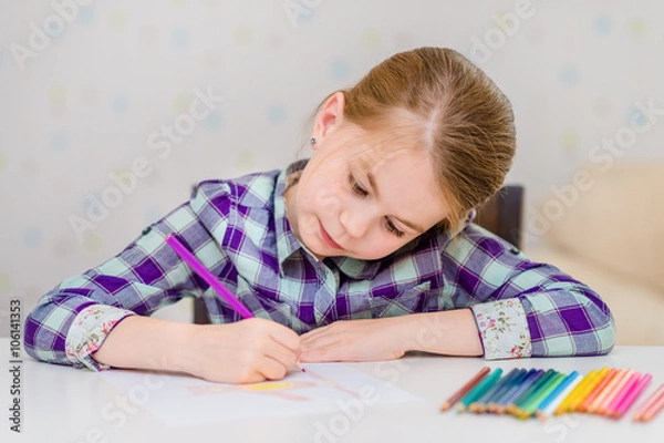 Fototapeta Beautiful pensive little girl with blond hair sitting at white table and drawing with multicolored pencils