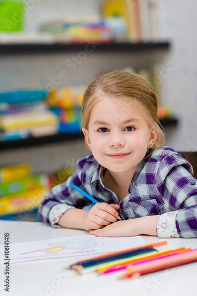 Obraz Lovely smiling little girl with blond hair sitting at white table with multicolored pencils and looking at camera