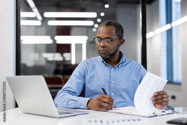 Fototapeta Mature African American businessman in modern office working on laptop, reviewing documents. Focused, professional, productive atmosphere, emphasizing business analysis, decision-making, and strategy