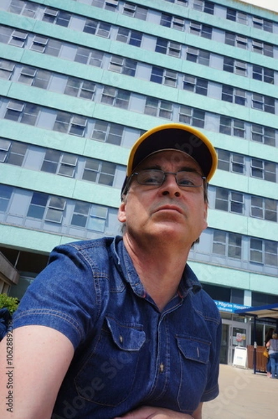 Fototapeta looking up at a mature man wearing a baseball cap and glasses in front of a multi-story building