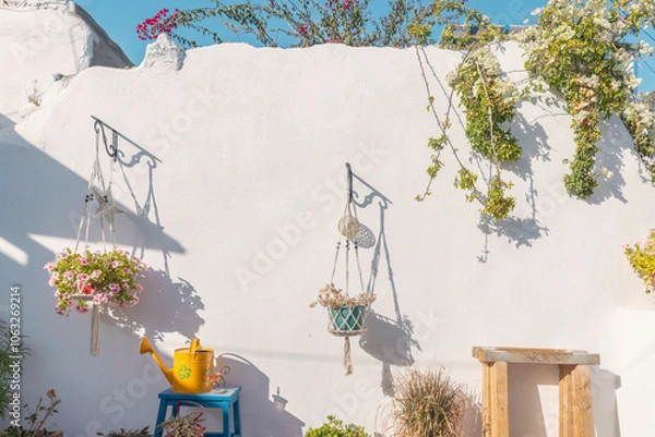 Obraz In a picturesque courtyard on Paros, vibrant hanging flower pots and a bright yellow watering can complement the whitewashed wall, creating a serene atmosphere beneath the sunny sky.