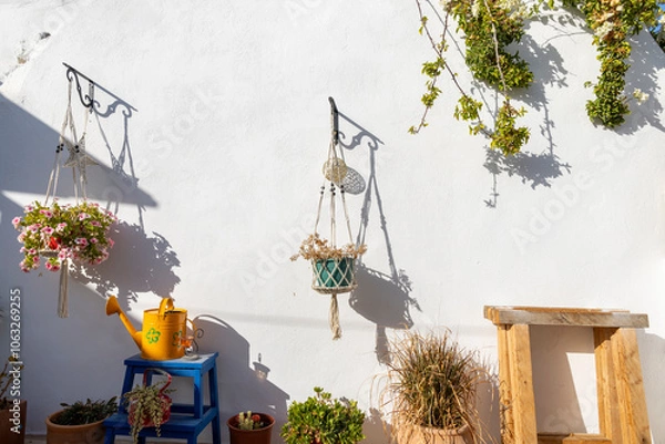 Obraz A sunlit wall in Paros features two hanging planters filled with flowers, accompanied by a yellow watering can, creating a welcoming outdoor atmosphere.