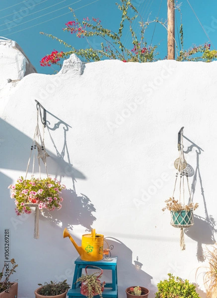 Obraz Hanging flower pots filled with blooms complement a whitewashed wall on Paros. A cheerful yellow watering can and wooden furniture enhance the garden ambiance in the Cyclades.