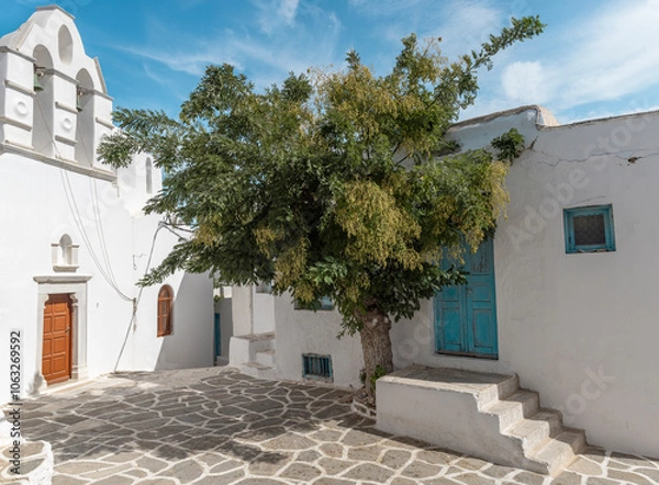 Obraz A picturesque alley in Paros features a traditional whitewashed house with bright blue doors, surrounded by quaint architecture and a lush tree casting a cool shade in the sun.