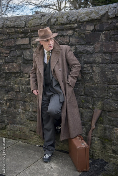 Fototapeta Vertical portrait image of a mature man dressed as a 1940s gangster character, with a suitcase and a shot gun.