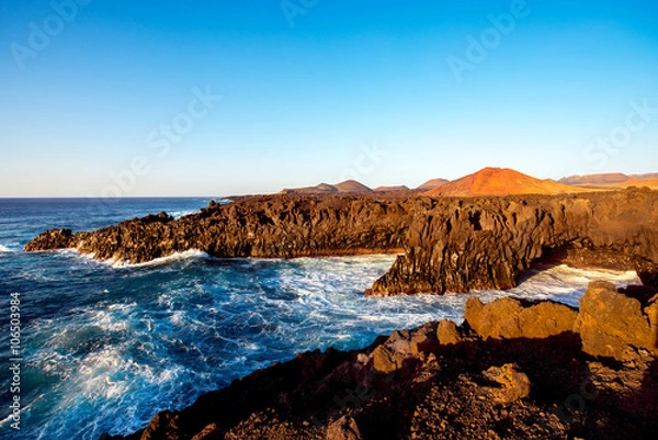 Obraz Los Hervideros rocky coast with wavy ocean and volcanos on the background on the sunset on Lanzarote island in Spain