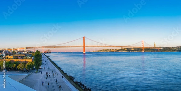 Obraz Panoramic view of 25 de abril bridge over tagus river in lisbon at sunset, with clear blue sky and christ the king statue in background, people enjoying waterfront promenade