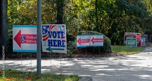 Fototapeta Vote Here signs directing voters to polling places, with secondary signs and candidate signs in soft focus in background.