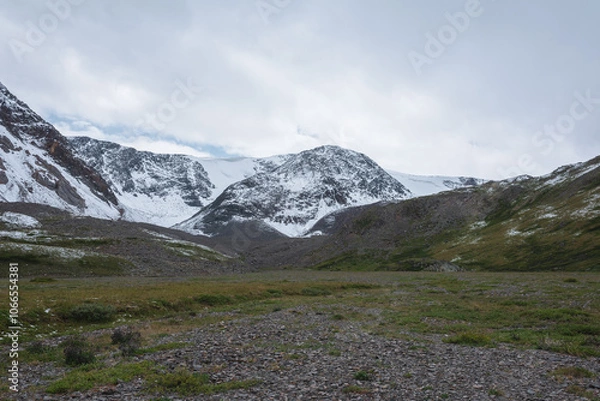 Fototapeta Dramatic cold landscape with green valley against snow covered large mountain range under rainy cloudy gray sky. Gloomy rainy weather in high mountains. Alpine view to big snowy mount in rain time.