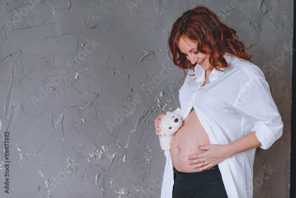 Fototapeta Pregnant woman in a white man's shirt, a studio portrait. Looking at the midsection. With a Teddy bear in front of him.