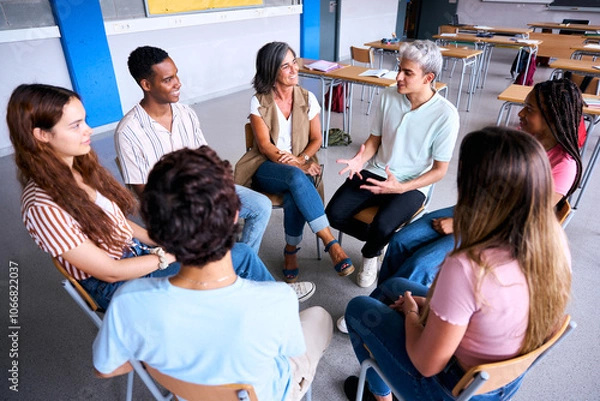 Fototapeta Diverse happy group of high school students sitting on chairs in circle interacting during lesson. Multiracial adolescents talking together to their classmates and female teacher in support class