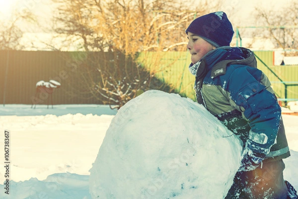 Fototapeta boy sculpt a snowman in the backyard of the house clear winter day. The boy rolls a snowball to build a snowman in the yard during a heavy snowfall. toned.
