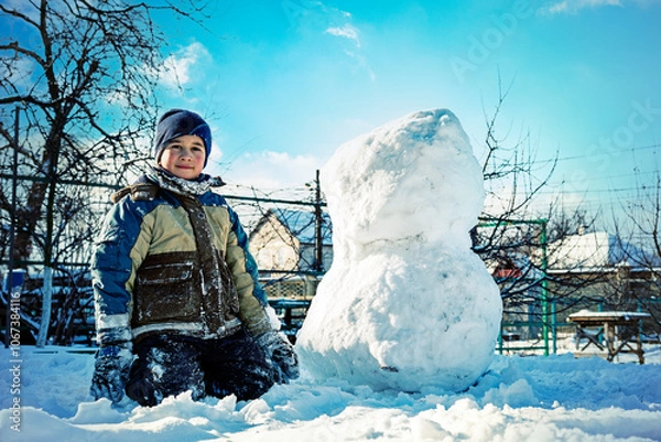 Fototapeta boy sculpt a snowman in the backyard of the house clear winter day. The boy rolls a snowball to build a snowman in the yard during a heavy snowfall. toned.