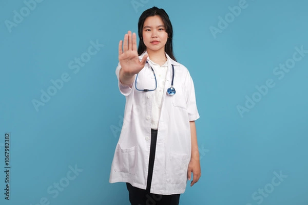 Obraz doctor holding a stethoscope. Angry young Asian woman doctor wearing uniform with a stethoscope frowning displeased, extend hand to show stopping, disagree, prohibit or forbid on blue background. Heal
